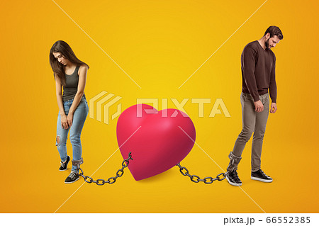 Young sad man and woman wearing casual clothes chained to pink heart on yellow background Young sad man and woman wearing casual clothes chained to pink heart on yellow background 66552385