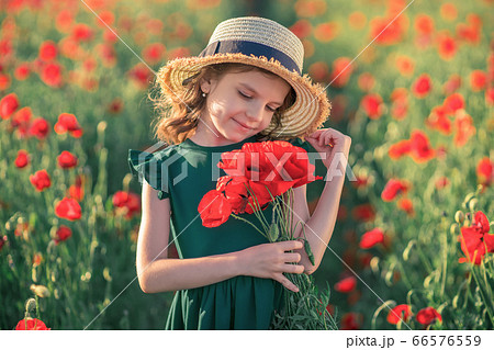 Cute little girl in green dress and straw hat with a bouquet of poppies posing at field of poppies in her hands on summer sunset 66576559
