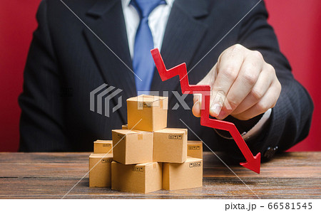 Businessman is holding a red arrow down above boxes stack pile. Business industrial production drop. Decrease freight transportation, volumes of delivery of products. Overproduction, scarcity of goods 66581545