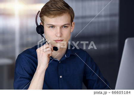 Young blond businessman using headset and computer in a darkened office, glare of light on the background. Startup business means working hard 66605184