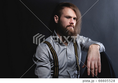 A portrait of an attractive brooding bearded man sitting against a wall looks nowhere and reflects on the dynocy and other problems. Studio portrait of stylish hipster 66605528