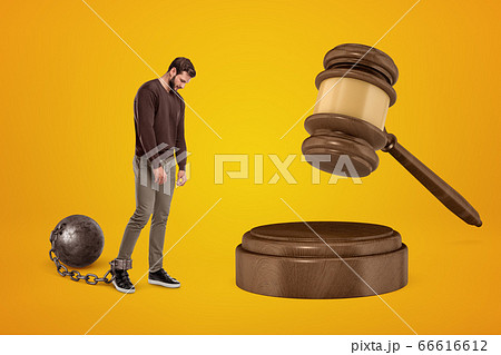 Distressed young man chained to shackle ball, looking down and walking slowly toward huge brown sound block with gavel on amber background. Distressed young man chained to shackle ball, looking down and walking slowly toward huge brown sound block with gavel on amber background. 66616612