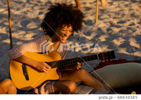 Young mixed race woman playing guitar and laughing on beach 66630365