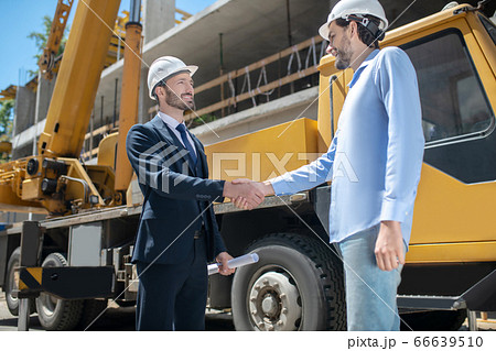 Building supervisor and foreman standing near crane, shaking hands with each other, happy to reach agreement Building supervisor and foreman standing near crane, shaking hands with each other, happy to reach agreement 66639510