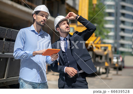 Foreman holding tablet, discussing something with building supervisor, touching his helmet with hand 66639528