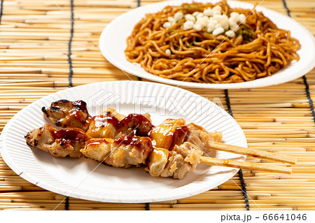 紙皿にのせられた焼き鳥 ねぎま たれ焼き 2本 写真奥には焼きそば 夏祭りの屋台メニューイメージ の写真素材