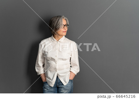 Beautiful mature lady in glasses stands hands in pockets looking at side. Elegant Gray-haired woman in whiite shirt on gray background. Copy space for text at right side Beautiful mature lady in glasses stands hands in pockets looking at side. Elegant Gray-haired woman in whiite shirt on gray background. Copy space for text at right side 66645216