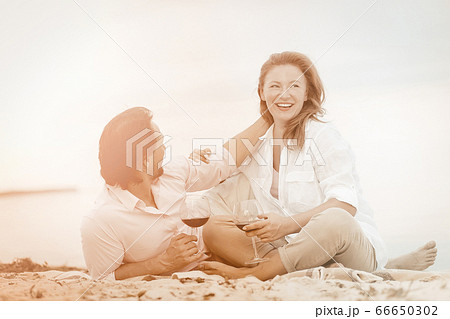 Happy couple enjoy chatting with each other, spending time together. Mature man and woman drink wine cuddling and laughing while sitting on sandy shore near water. Vacation by sea. Tinted image Happy couple enjoy chatting with each other, spending time together. Mature man and woman drink wine cuddling and laughing while sitting on sandy shore near water. Vacation by sea. Tinted image 66650302