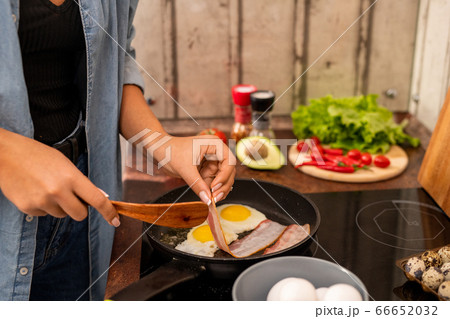 Young female standing by electric stove and putting slice of bacon on frying pan Young female standing by electric stove and putting slice of bacon on frying pan 66652032