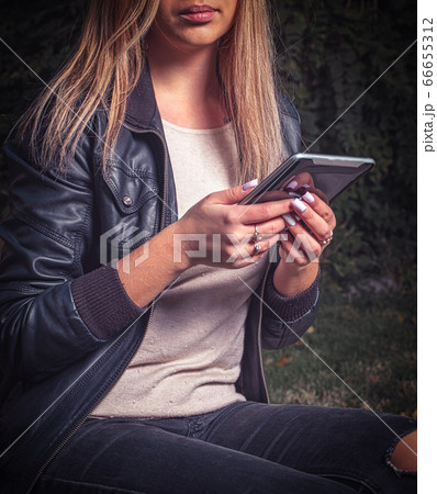 A female student in a black leather jacket and ripped jeans, she is engaged on her tablet computer in the park A female student in a black leather jacket and ripped jeans, she is engaged on her tablet computer in the park 66655312