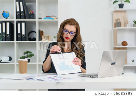 Experienced successful business lady in stylish clothes and glasses talking with her business partner by video call and shows report with graphs. 66685398