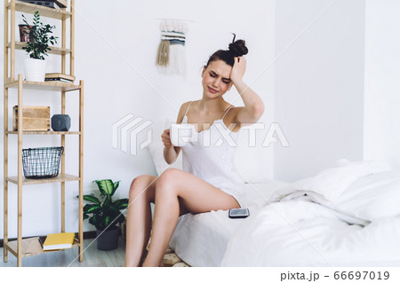 Upset attractive woman sitting in bed with coffee cup and having headache 66697019