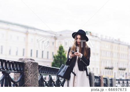 Portrait of a stylish hipster girl dressed in cool clothes posing on the street while holding cell telephone 66697673