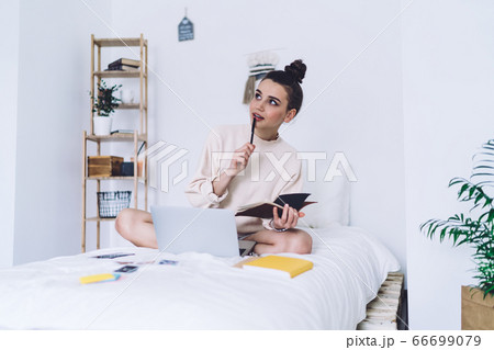 Creative young wistful woman remote worker sitting on cozy bed and analyzing ideas 66699079