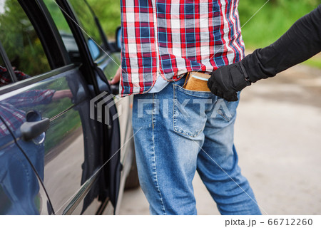 The thief in black clothes and gloves stealing a wallet with money from pocket near the car. Pickpocketing on the street during daytime. 66712260