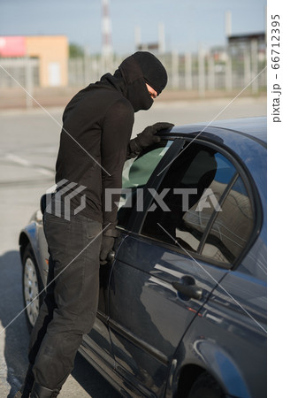 An offender in black clothes and mask is trying to steal a car in a parking lot. Stop criminality. 66712395