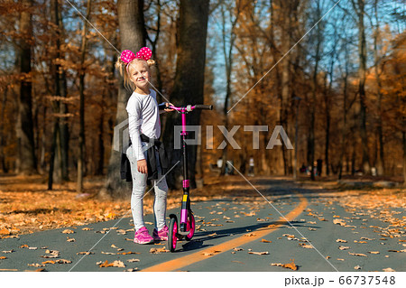 Cute adorable little caucasian school girl portrait having fun riding pink kick scooter bike in golden autumn city park or forest outside. Happy childhood. Kids outdoor sport activities 66737548