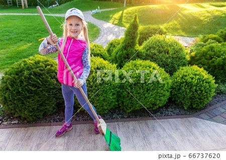 Cute adorable little caucasian girl hepling parents sweeping yard with broom on warm bright autumn evening outdoors. Happy young playful kid making housework in ornamental backyard garden 66737620