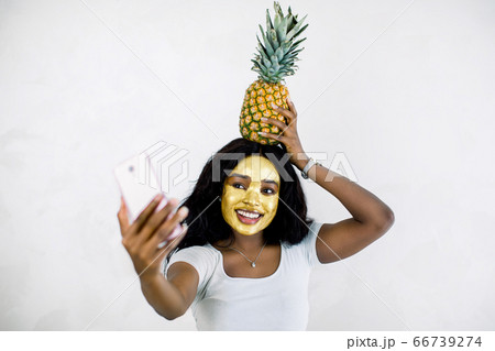 Attractive African girl with golden peel off mask, pineapple on her head, taking selfie photo with smartphone, enjoying relaxation and beauty treatment. Woman with facial mask making selfie Attractive African girl with golden peel off mask, pineapple on her head, taking selfie photo with smartphone, enjoying relaxation and beauty treatment. Woman with facial mask making selfie 66739274