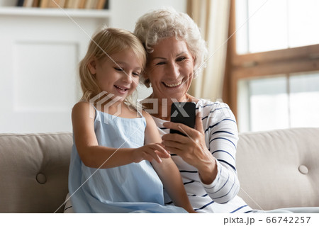Little granddaughter aged grandmother having fun at home using smartphone Little granddaughter aged grandmother having fun at home using smartphone 66742257