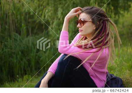 Outdoors portrait of a girl with dreadlocks wearing pink sweater and sunglasses in a city park during a sunset, be free concept. 66742424