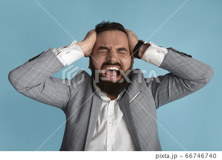 Millennial businessman holding his head and screaming in anger on blue background Millennial businessman holding his head and screaming in anger on blue background 66746740