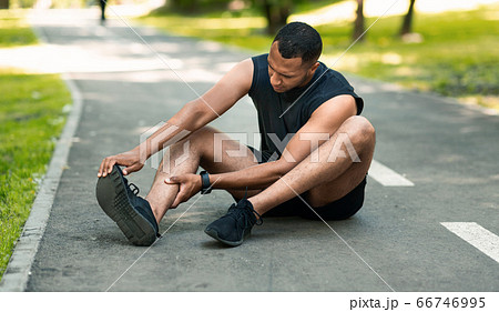 Sports injury. African American runner sitting on jogging track and feeling pain in his ankle Sports injury. African American runner sitting on jogging track and feeling pain in his ankle 66746995