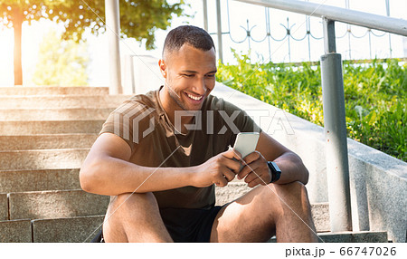 Happy black guy in sportswear relaxing after his workout on stairs and browsing internet on smartphone, outdoors Happy black guy in sportswear relaxing after his workout on stairs and browsing internet on smartphone, outdoors 66747026