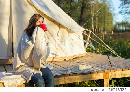 Young woman wraps blanket over herself while drinking coffee near canvas tent in the morning 66748522