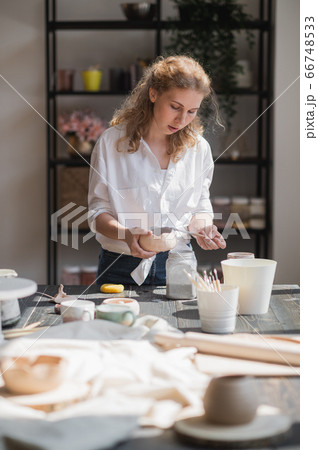Female potter glazing raw unburned ceramic cup using brush. Earthenware mug putting colorant on pottery workshop. 66748533
