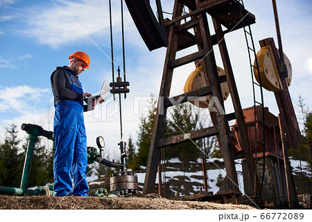 Oil engineer with a laptop standing next to an oil rig making notes in his computer Oil engineer with a laptop standing next to an oil rig making notes in his computer 66772089