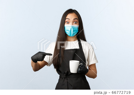 Covid-19, social distancing, small coffee shop business and preventing virus concept. Excited asian barista, female cafe employee in medical mask and gloves pointing at mug with coffee Covid-19, social distancing, small coffee shop business and preventing virus concept. Excited asian barista, female cafe employee in medical mask and gloves pointing at mug with coffee 66772719