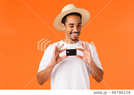 Relaxed and cheeky cute african-american man in white t-shirt, summer hat, wink camera assured and pleased, recommend bank service, holding credit card, standing orange background 66786226