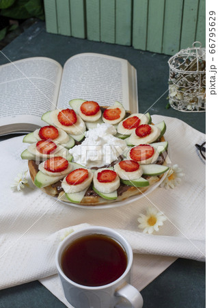 Waffles with banana and strawberry served with a cup of tea on a white tablecloth, a book around 66795629