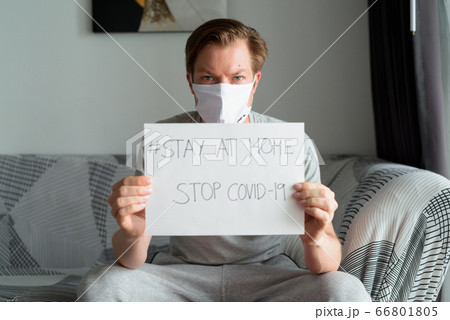 Young man with mask showing Stay At Home sign in the living room while under quarantine Young man with mask showing Stay At Home sign in the living room while under quarantine 66801805