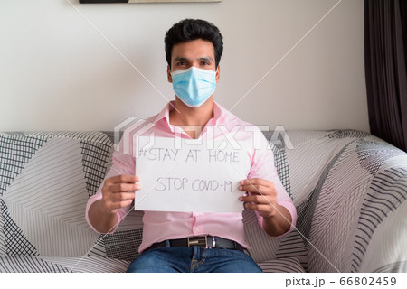 Young Indian businessman with mask showing Stay At Home sign while sitting in the living room under 66802459