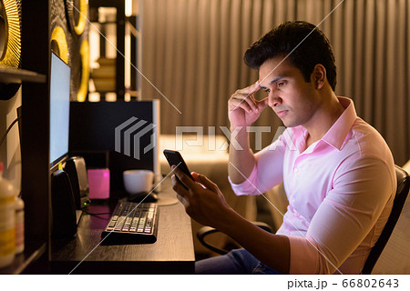 Stressed young Indian businessman using phone while working overtime at home late at night Stressed young Indian businessman using phone while working overtime at home late at night 66802643