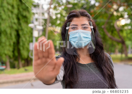 Young Indian woman with mask and face shield showing stop gesture outdoors 66803235