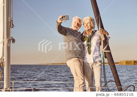 Happy senior couple standing on the side of sail boat or yacht deck floating in sea. Man and woman smiling, while taking selfie using smartphone 66805216