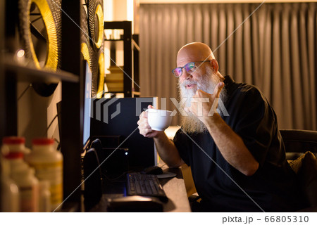 Happy mature bald bearded man drinking coffee while video calling at work from home late at night Happy mature bald bearded man drinking coffee while video calling at work from home late at night 66805310