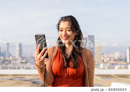 Happy young beautiful Indian woman using phone against view of the city Happy young beautiful Indian woman using phone against view of the city 66806004