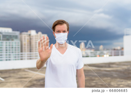 Young man with mask showing stop gesture against view of the city during stormy weather outdoors Young man with mask showing stop gesture against view of the city during stormy weather outdoors 66806685