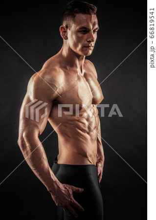 Fitness in gym, sport and healthy lifestyle concept. Handsome athletic man showing his trained body on dark background. Bodybuilder male model standing with crossed hands and tight muscles. 66819541
