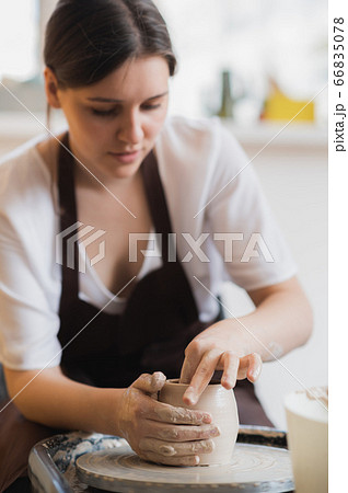 Closeup of young female potter with sponge keeping it close to rotating clay pot while shaping form of pot during work Closeup of young female potter with sponge keeping it close to rotating clay pot while shaping form of pot during work 66835078