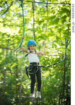 Rope adventure in the forest - a little girl standing on the bridge with full insurance and having an entertainment - looking in the camera Rope adventure in the forest - a little girl standing on the bridge with full insurance and having an entertainment - looking in the camera 66838583