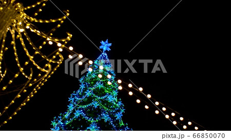 View of Christmas tree decorated large luminous multi-pointed blue stars and yellow balls through garland of yellow lights encircling city square View of Christmas tree decorated large luminous multi-pointed blue stars and yellow balls through garland of yellow lights encircling city square 66850070
