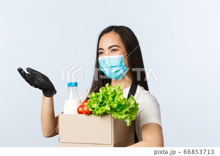 Covid-19, grocery store, employment, small business and virus concept. Smiling cashier, asian shop worker in medical mask stretch hand to except payment in cash, holding packed groceries order 66853713