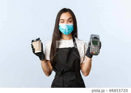 Coffee shop, coronavirus, social distancing and contactless payment concept. Cheeky cute asian waitress, barista in medical mask and gloves, wink as handing coffee order and paying terminal Coffee shop, coronavirus, social distancing and contactless payment concept. Cheeky cute asian waitress, barista in medical mask and gloves, wink as handing coffee order and paying terminal 66853720
