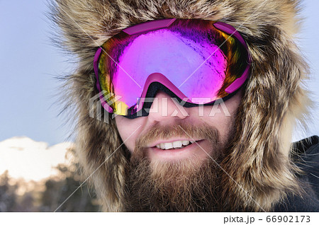 Close-up portrait of a bearded happy snowboarder skier in a ski mask with goggles and a fur big old-school hat on a background of a winter snowy mountains 66902173