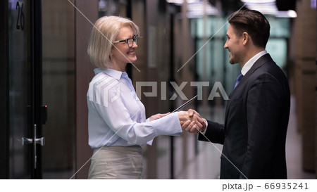 Older businesswoman and businessman shaking hands standing in office corridor Older businesswoman and businessman shaking hands standing in office corridor 66935241